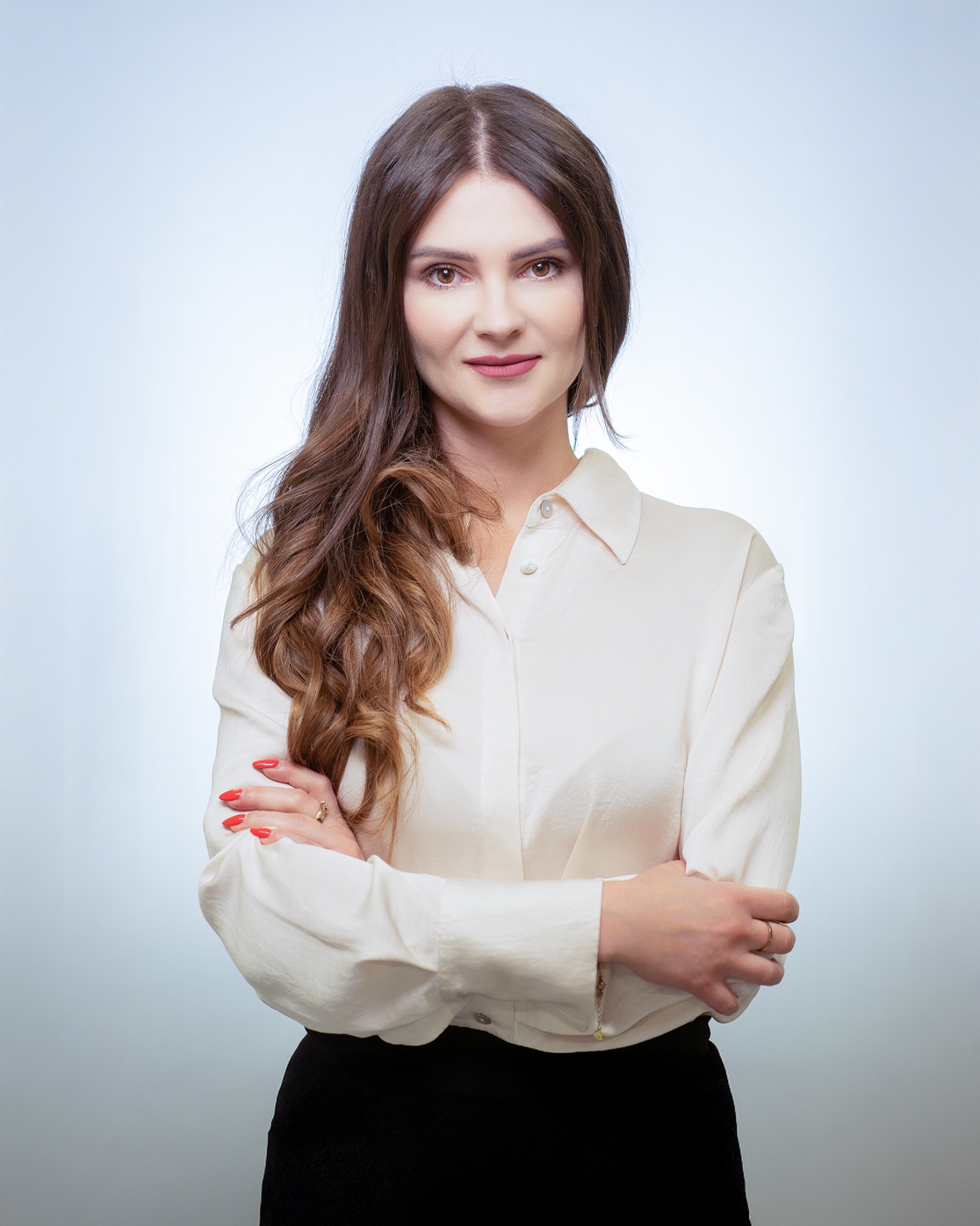 Portret uśmiechniętej kobiety w białej koszuli z podwiniętymi rękawami i czarnej spódnicy, skrzyżowane ramiona, czerwone paznokcie i złoty pierścionek na palcu, brązowe włosy opadające na ramię...