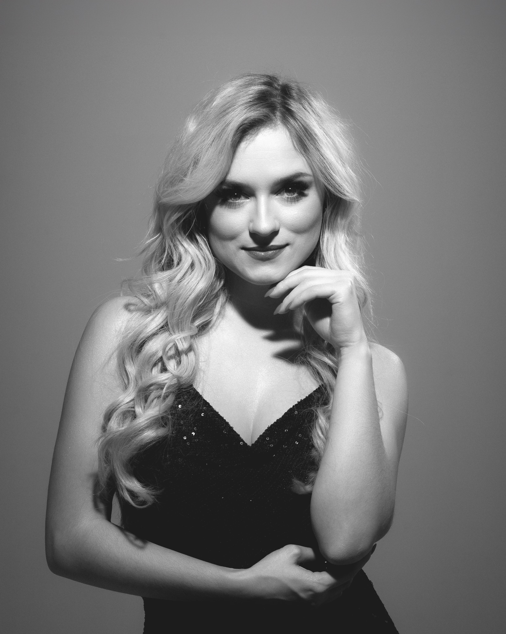 Portret kobiety z blond włosami, opartą brodą na dłoni, w czarnej, cekinowej sukience na szarym tle. Zdjęcie czarno-białe.