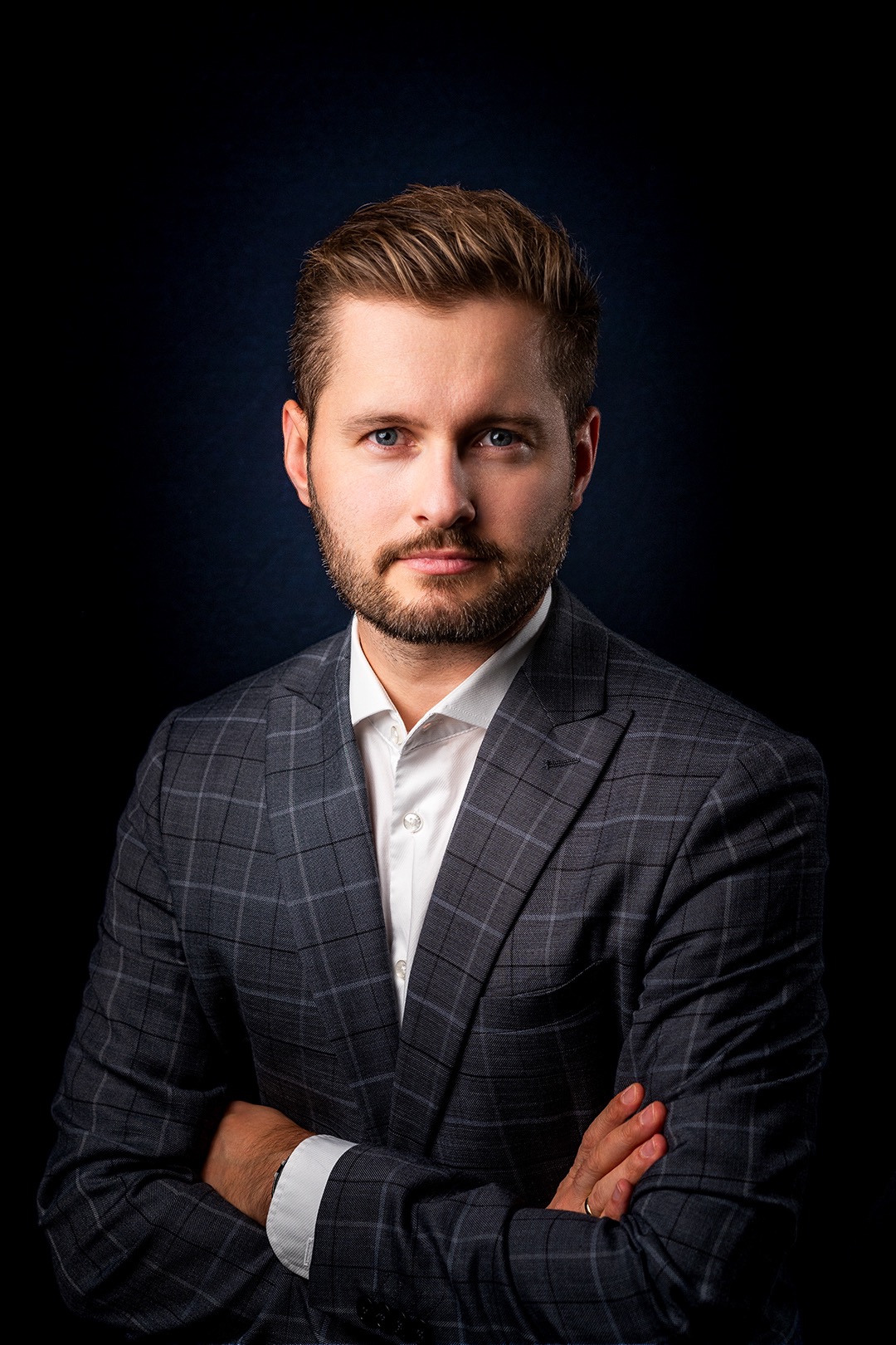 Portret biznesowy mężczyzny w szarej marynarce w kratę i białej koszuli, z założonymi rękami na ciemnym tle.