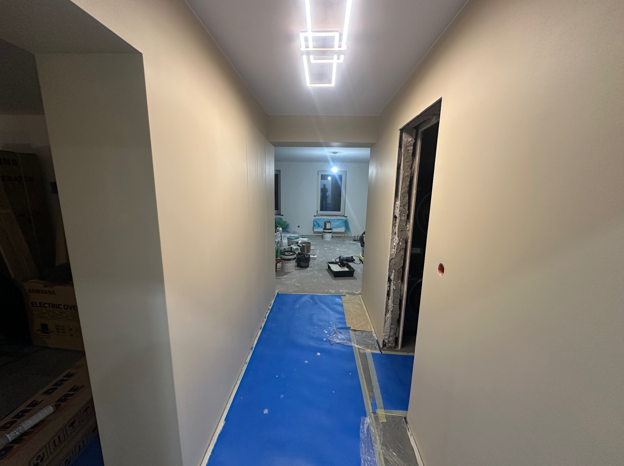 Korytarz w trakcie remontu: świeżo pomalowane ściany w odcieniach beżu, zabezpieczona podłoga folią w kolorze niebieskim, widoczne puszki po farbach i narzędzia malarskie w oddali, nowoczesne...