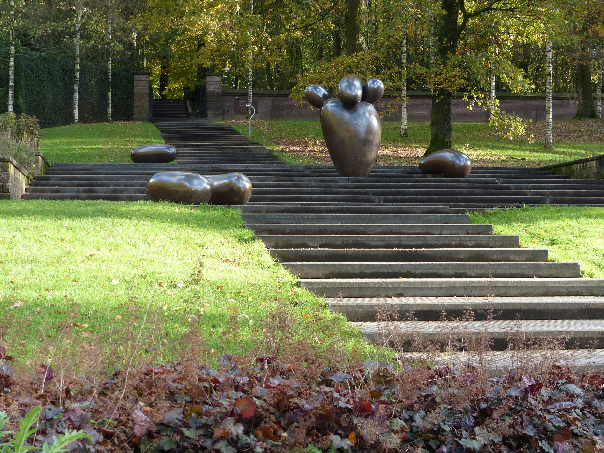 Schody z rzeźbami w parku, widok na wzniesienie z zielonym trawnikiem i drzewami w tle.