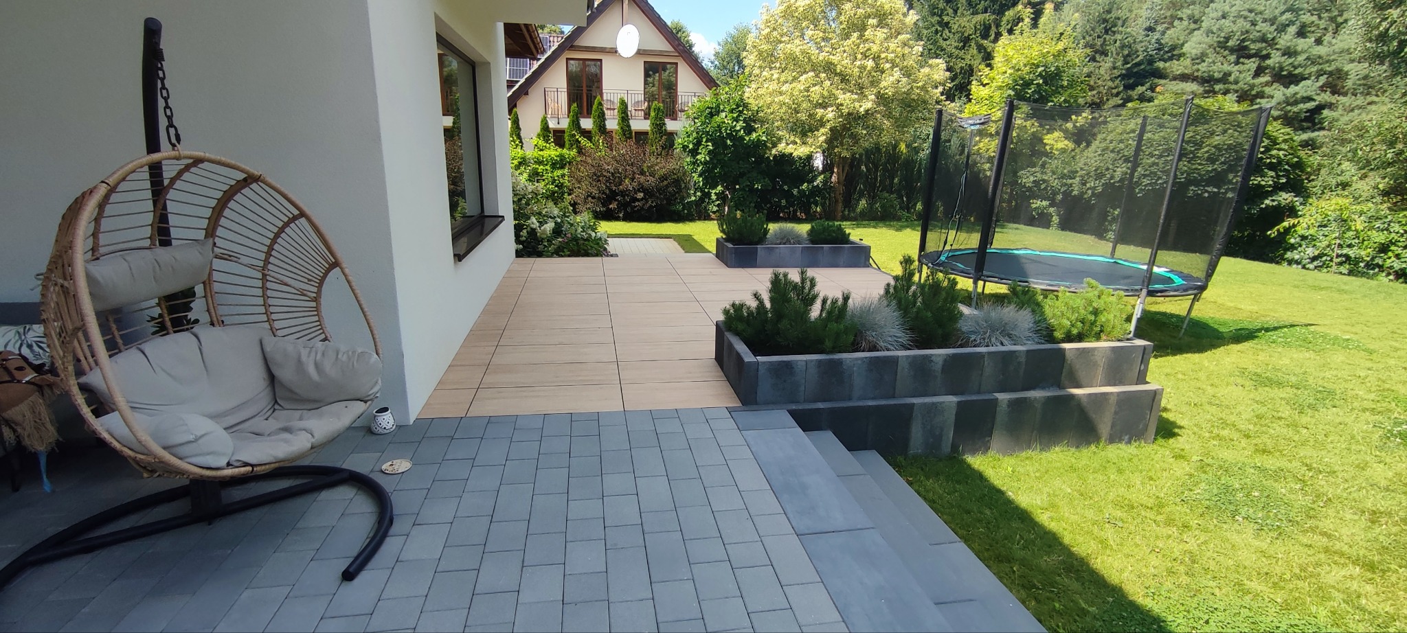 Nowoczesny ogród z huśtawką kokon, tarasem z płytek i trampoliną. Donice z roślinami ozdobnymi podkreślają minimalistyczny charakter przestrzeni.