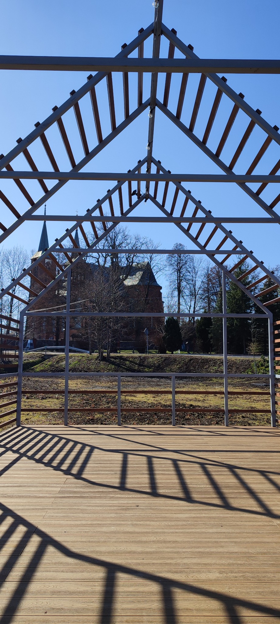 Drewniany taras z metalową pergolą w kształcie dachu, rzucającą cienie na deski. W tle budynek z wieżą i drzewa pod błękitnym niebem.