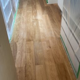 Mateusz Kurek - Świeżo ułożona, lakierowana podłoga z paneli drewnopodobnych w jasnym odcieniu dębu, zabezpieczona folią i taśmą malarską przed dalszymi pracami wykończeniowymi.