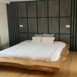 Sypialnia z łóżkiem na drewnianej platformie przypominającej plaster pnia drzewa, ściana za łóżkiem w odcieniach szarości z zielonymi listwami tworzącymi geometryczny wzór, drewniana podłoga.