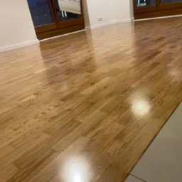 Błyszcząca, świeżo ułożona podłoga z parkietu w jasnym odcieniu drewna, widoczne okna z żaluzjami w tle, listwy przypodłogowe.