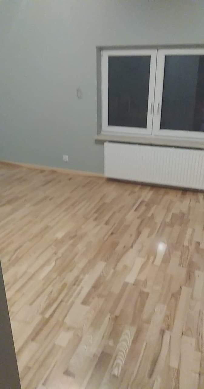 Jasny, drewniany parkiet ułożony w nowym pomieszczeniu z szarą ścianą i białym grzejnikiem pod oknem.