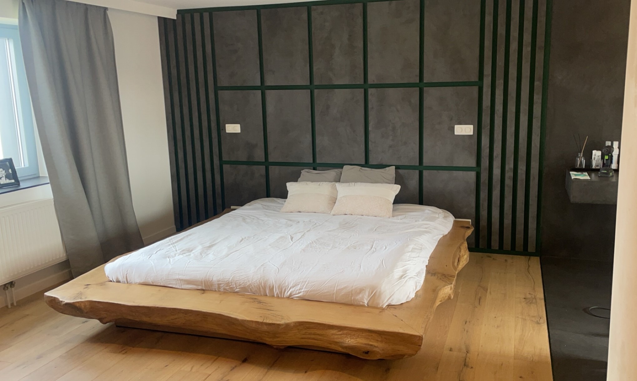 Sypialnia z łóżkiem na drewnianej platformie przypominającej plaster pnia drzewa, ściana za łóżkiem w odcieniach szarości z zielonymi listwami tworzącymi geometryczny wzór, drewniana podłoga.