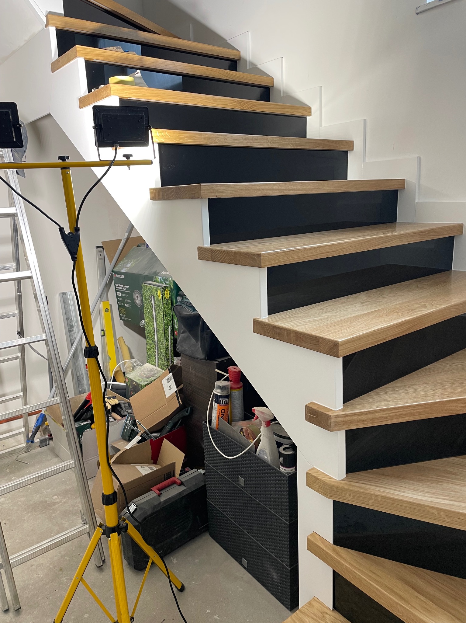 Nowoczesne schody z drewnianymi stopniami, czarnymi podstopnicami i białymi bokami, oświetlone lampą budowlaną, w tle widoczny nieporządek i drabina.