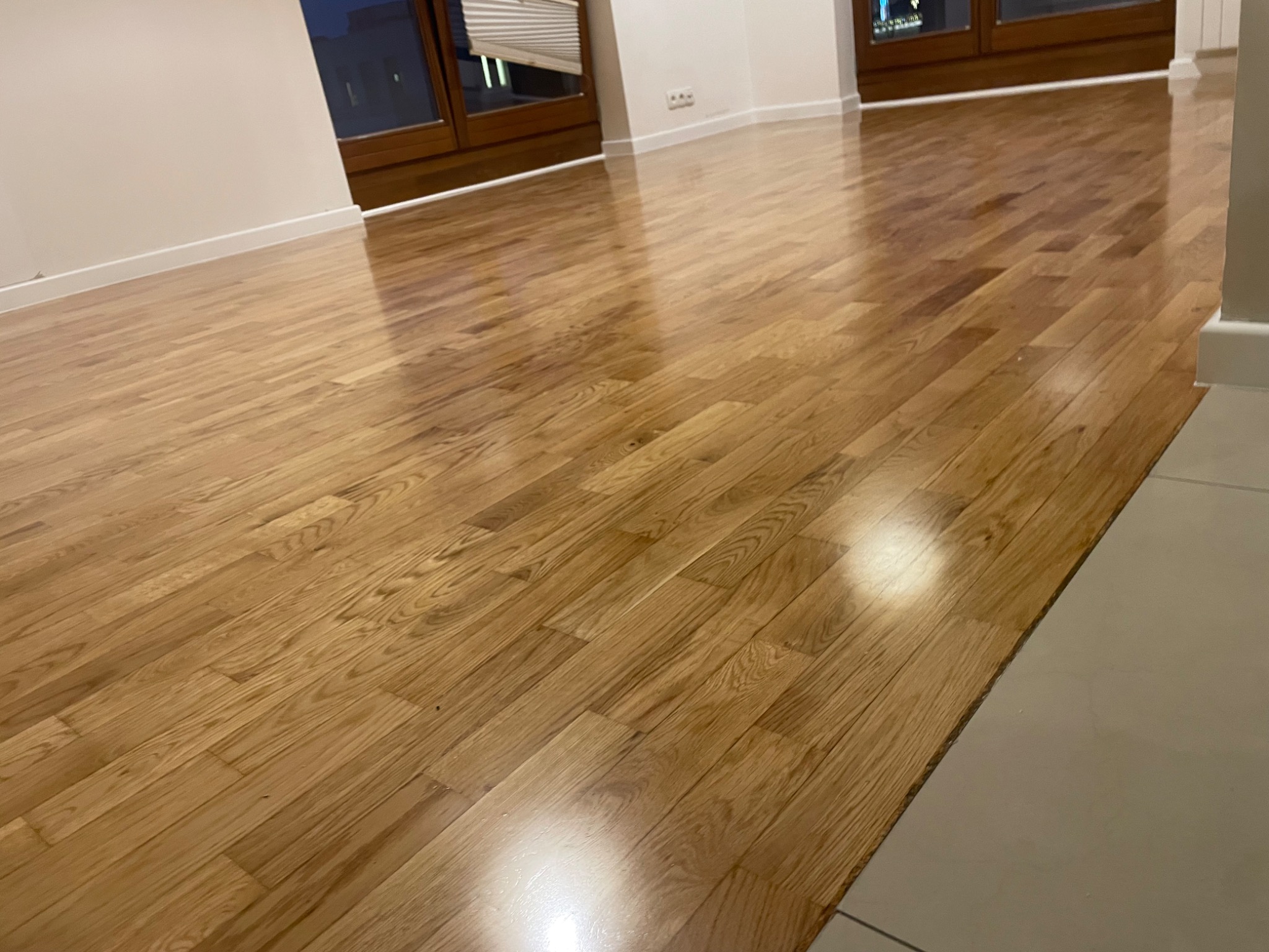 Błyszcząca, świeżo ułożona podłoga z parkietu w jasnym odcieniu drewna, widoczne okna z żaluzjami w tle, listwy przypodłogowe.