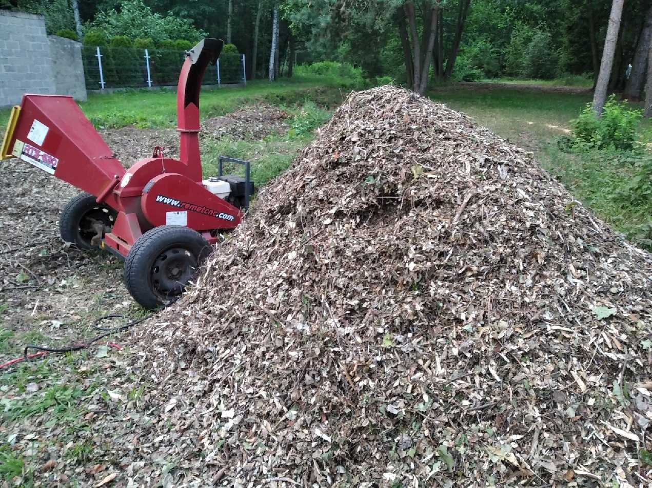 Czerwony rębacz do gałęzi obok pryzmy zrębków na trawie. Widok na zieleń ogrodu i drzewa w tle. Na maszynie widoczny adres strony internetowej.