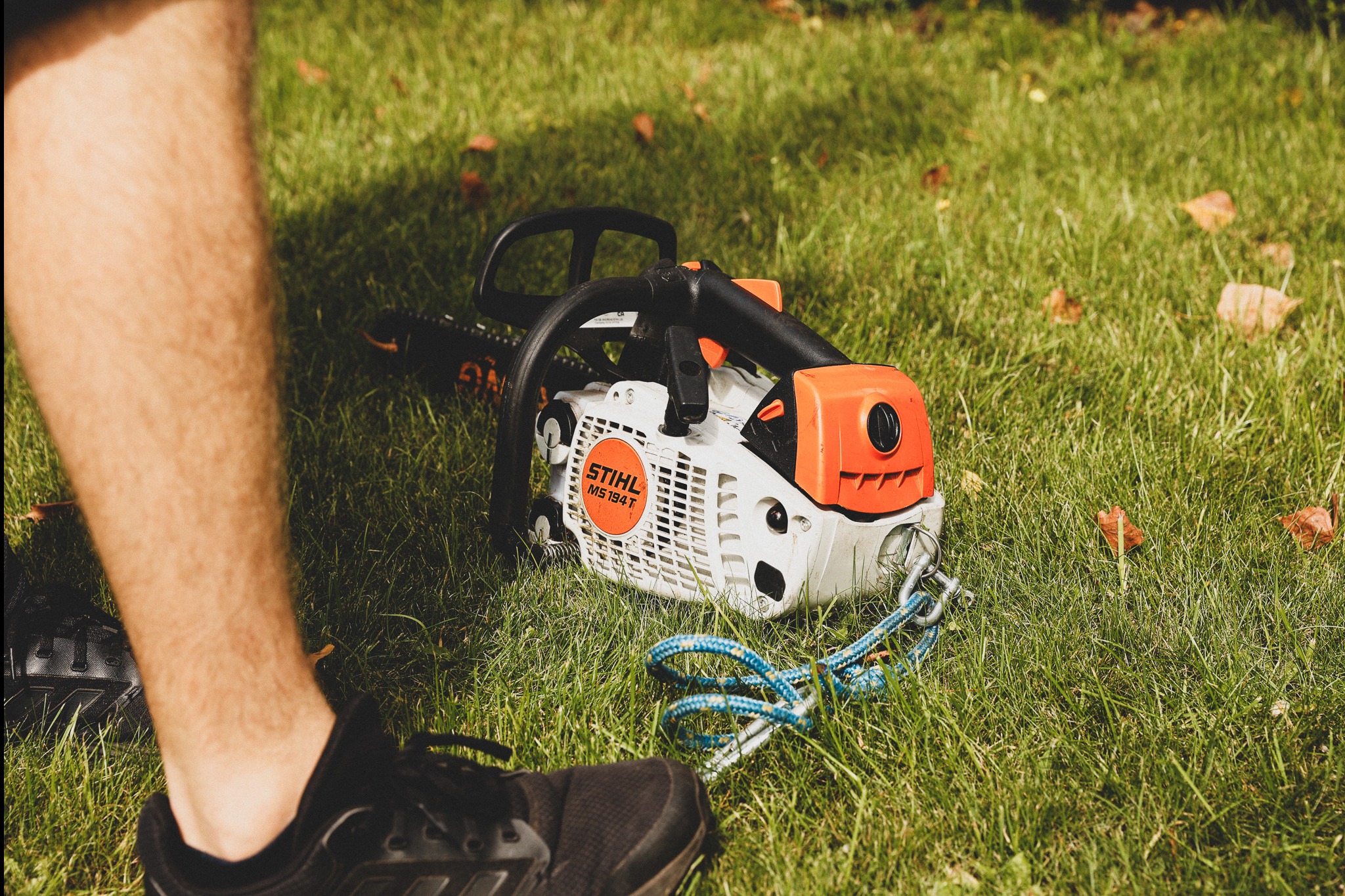 Piła spalinowa Stihl MS 194 T na trawie z nogą i butem w kadrze. Sprzęt ogrodniczy w zbliżeniu, jesienne liście na trawniku.