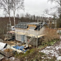 NILAMI SPÓŁKA Z OGRANICZONĄ ODPOWIEDZIALNOŚCIĄ - Budowa domu szkieletowego w trakcie realizacji, widoczne rusztowania, elementy konstrukcyjne, papa na dachu, ocieplenie ścian oraz materiały budowlane na działce w pochmurny dzień.