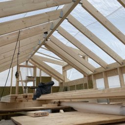 NILAMI SPÓŁKA Z OGRANICZONĄ ODPOWIEDZIALNOŚCIĄ - Wnętrze domu w trakcie budowy, widoczne drewniane elementy konstrukcyjne dachu i ścian, ocieplenie z wełny mineralnej, narzędzia i materiały budowlane na podłodze, całość oświetlona naturalnym...