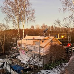 NILAMI SPÓŁKA Z OGRANICZONĄ ODPOWIEDZIALNOŚCIĄ - Budowa domu szkieletowego w trakcie realizacji, widoczne rusztowania, izolacja ścian, drewniana konstrukcja dachu i pracownik w czapce na dachu, drzewa bez liści w tle.