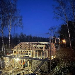 NILAMI SPÓŁKA Z OGRANICZONĄ ODPOWIEDZIALNOŚCIĄ - Konstrukcja domu szkieletowego w trakcie budowy, widoczna drewniana rama, rusztowania, wieczorne oświetlenie, błękitne niebo w tle.