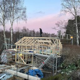 NILAMI SPÓŁKA Z OGRANICZONĄ ODPOWIEDZIALNOŚCIĄ - Wznoszenie konstrukcji domu szkieletowego na fundamencie z bloczków betonowych, widoczne rusztowania, pracownik na dachu i malownicze, różowe niebo o zmierzchu.