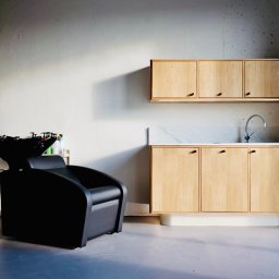 NILAMI SPÓŁKA Z OGRANICZONĄ ODPOWIEDZIALNOŚCIĄ - Nowoczesne wnętrze salonu fryzjerskiego: czarny fotel z umywalką, drewniane szafki z marmurowym blatem i chromowaną baterią, minimalistyczny grzejnik na ścianie o surowym wykończeniu.