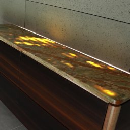 NILAMI SPÓŁKA Z OGRANICZONĄ ODPOWIEDZIALNOŚCIĄ - Komoda z ciemnego drewna z podświetlanym blatem z zielono-brązowego kamienia na tle szarej ściany z płytek.