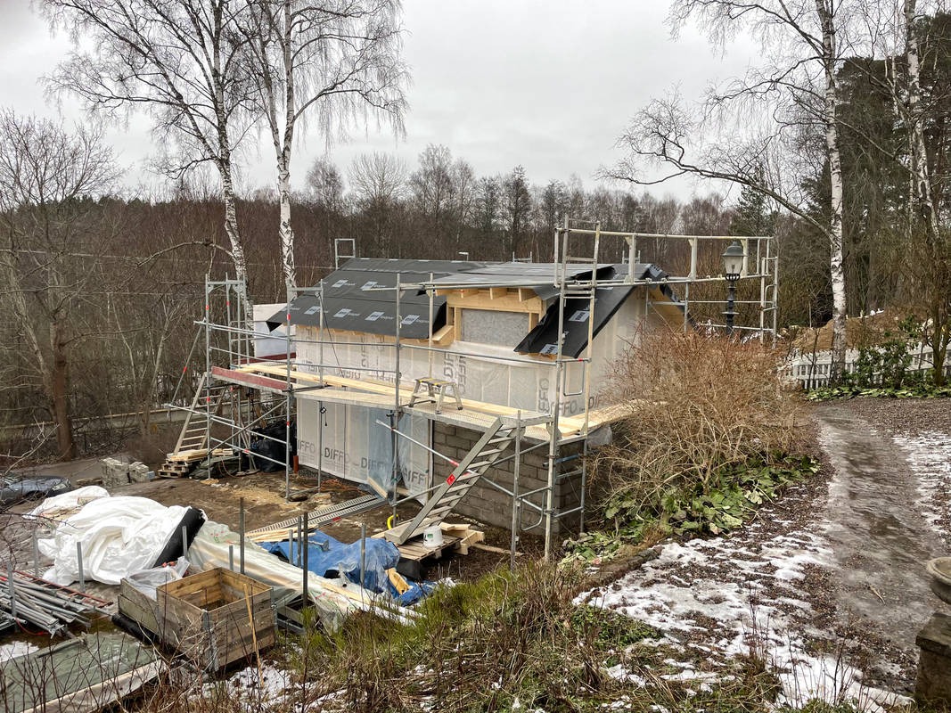 Budowa domu szkieletowego w trakcie realizacji, widoczne rusztowania, elementy konstrukcyjne, papa na dachu, ocieplenie ścian oraz materiały budowlane na działce w pochmurny dzień.