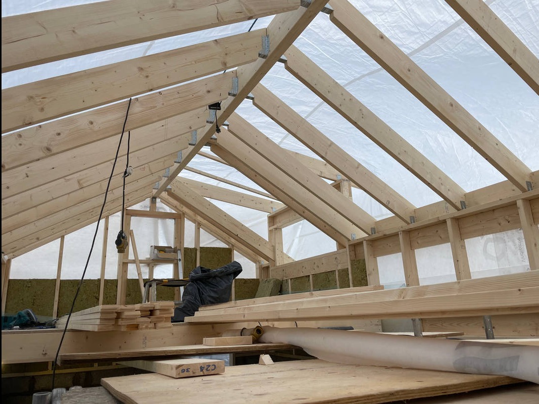 Wnętrze domu w trakcie budowy, widoczne drewniane elementy konstrukcyjne dachu i ścian, ocieplenie z wełny mineralnej, narzędzia i materiały budowlane na podłodze, całość oświetlona naturalnym...