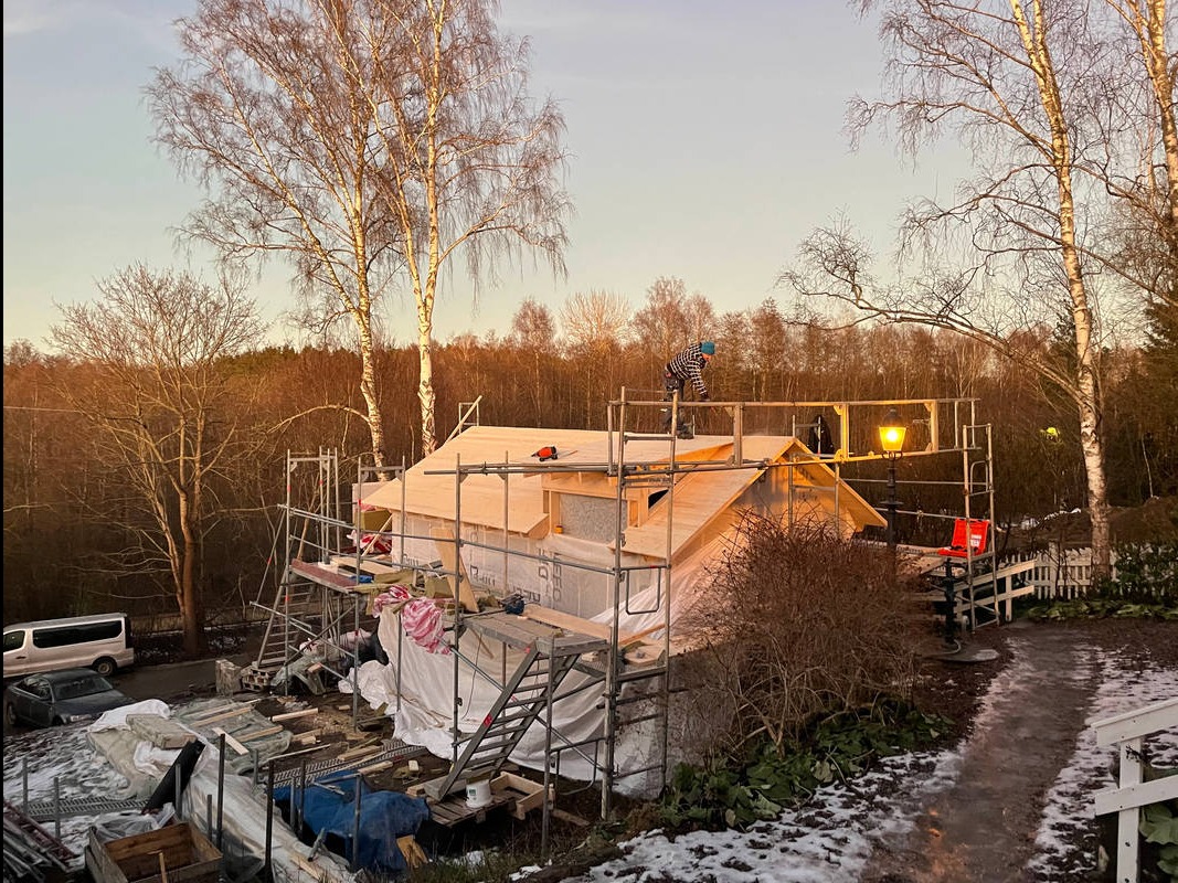 Budowa domu szkieletowego w trakcie realizacji, widoczne rusztowania, izolacja ścian, drewniana konstrukcja dachu i pracownik w czapce na dachu, drzewa bez liści w tle.
