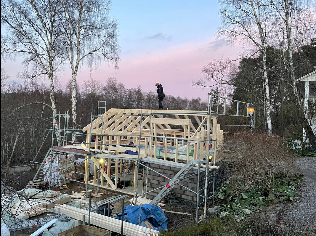 Wznoszenie konstrukcji domu szkieletowego na fundamencie z bloczków betonowych, widoczne rusztowania, pracownik na dachu i malownicze, różowe niebo o zmierzchu.