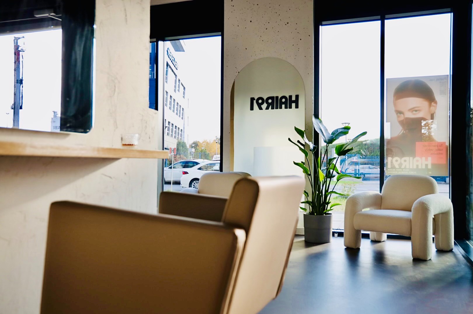 Wnętrze salonu fryzjerskiego z beżowymi fotelami, rośliną w szarej doniczce, plakatem reklamowym i logo na ścianie, widok na ulicę z białymi samochodami przez okno.