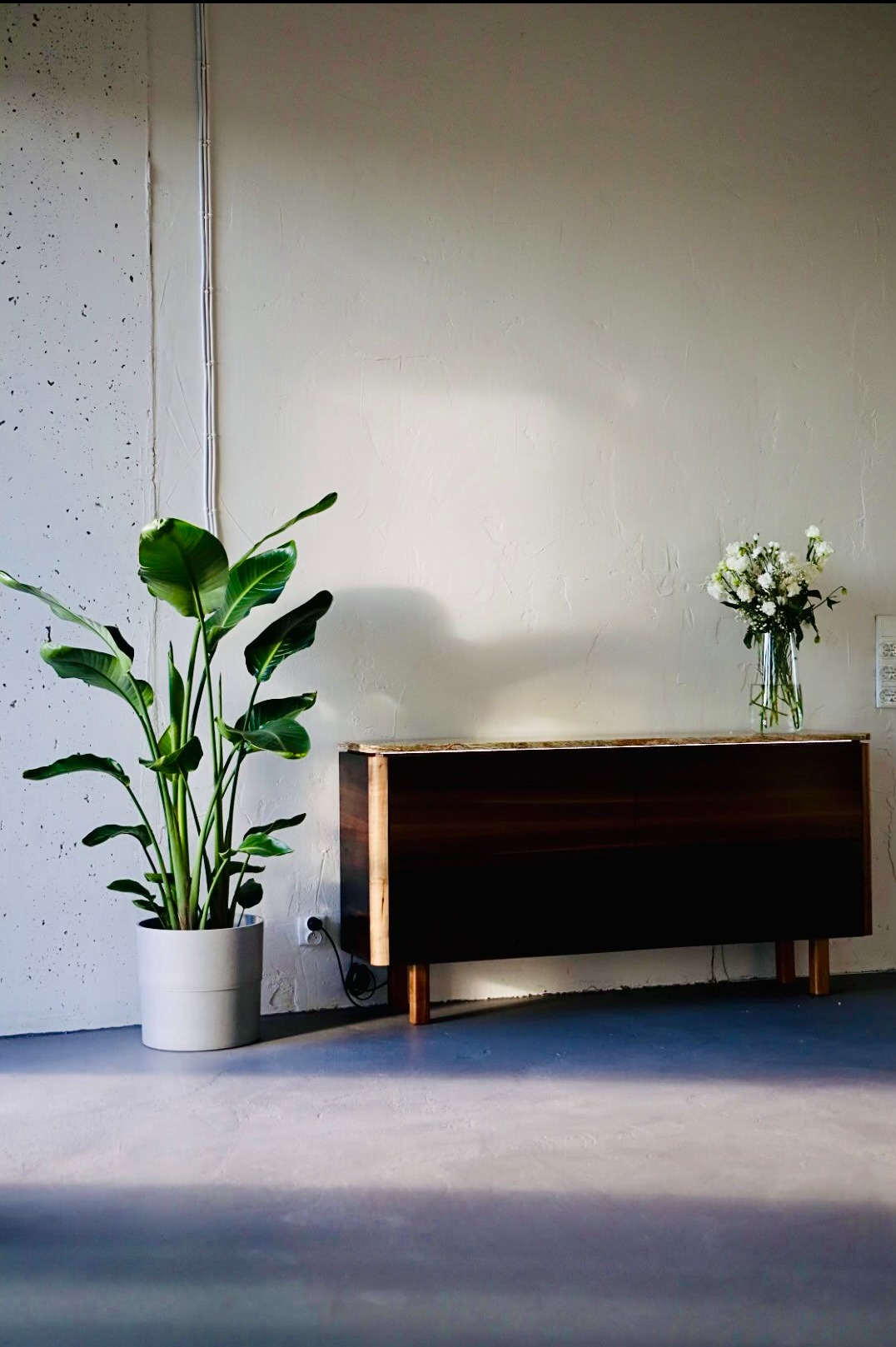 Minimalistyczne wnętrze z ciemnym drewnianym kredensem na niskich nóżkach, białym bukietem w szklanym wazonie, strelicją w szarej donicy, szarą podłogą i białymi ścianami o różnej fakturze.