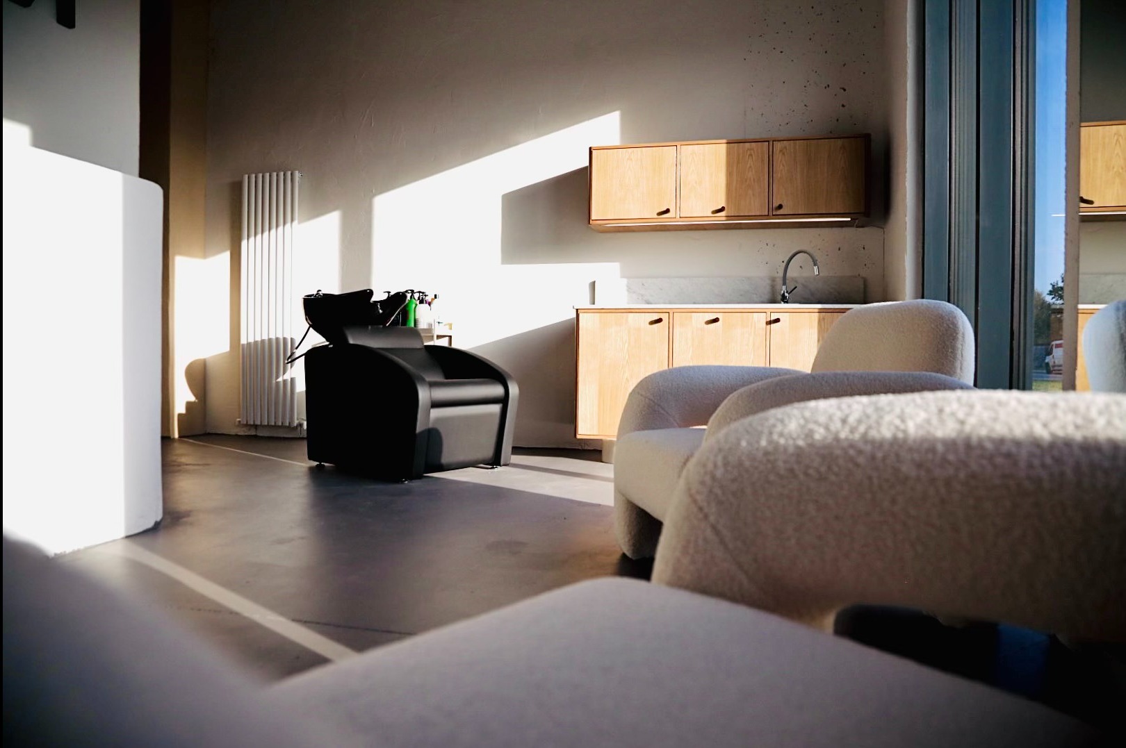 Nowoczesne wnętrze salonu fryzjerskiego z czarnym fotelem do mycia włosów, jasnymi szafkami z umywalką i białymi fotelami dla klientów, oświetlone naturalnym światłem wpadającym przez okno.