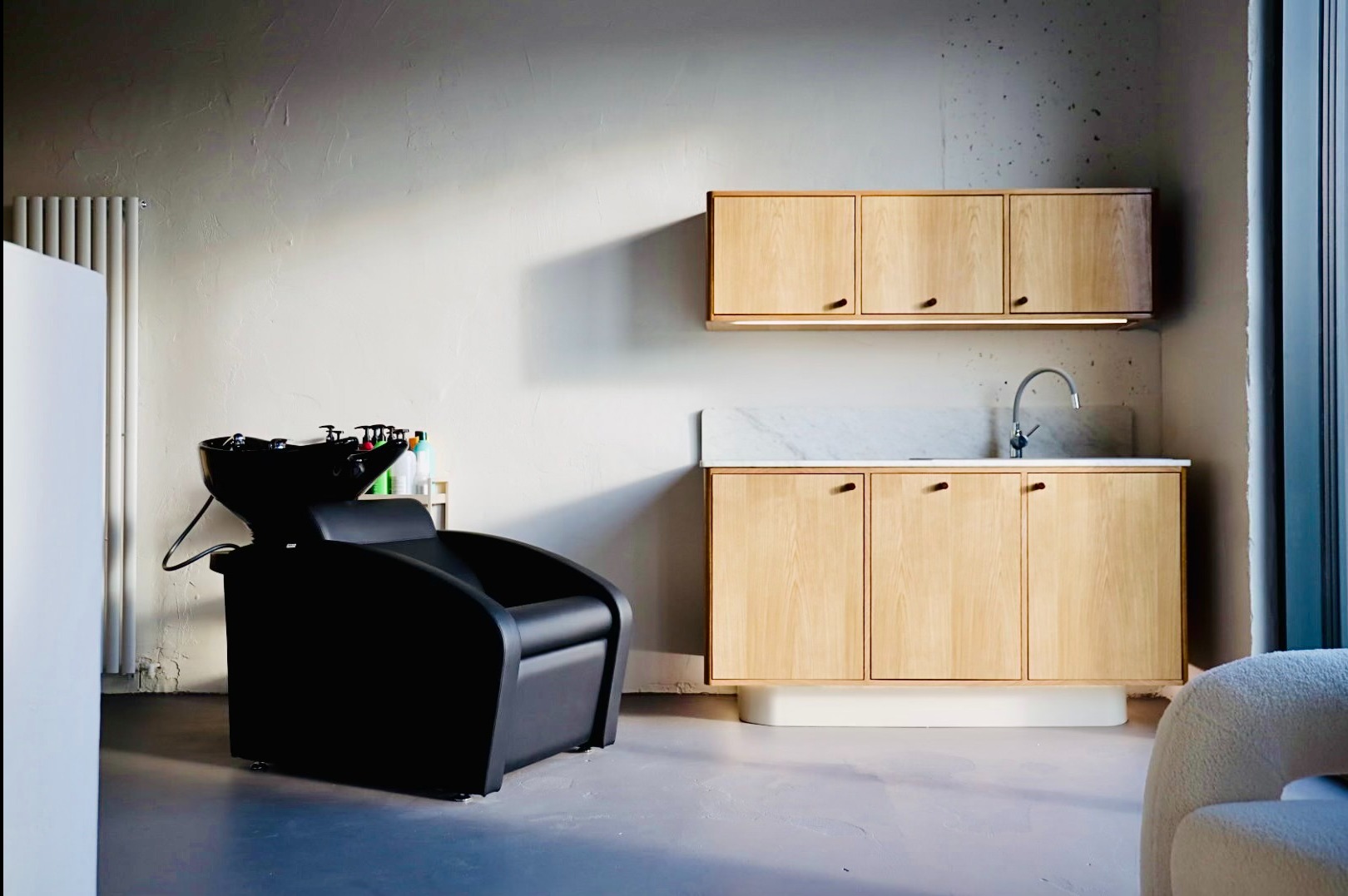 Nowoczesne wnętrze salonu fryzjerskiego: czarny fotel z umywalką, drewniane szafki z marmurowym blatem i chromowaną baterią, minimalistyczny grzejnik na ścianie o surowym wykończeniu.