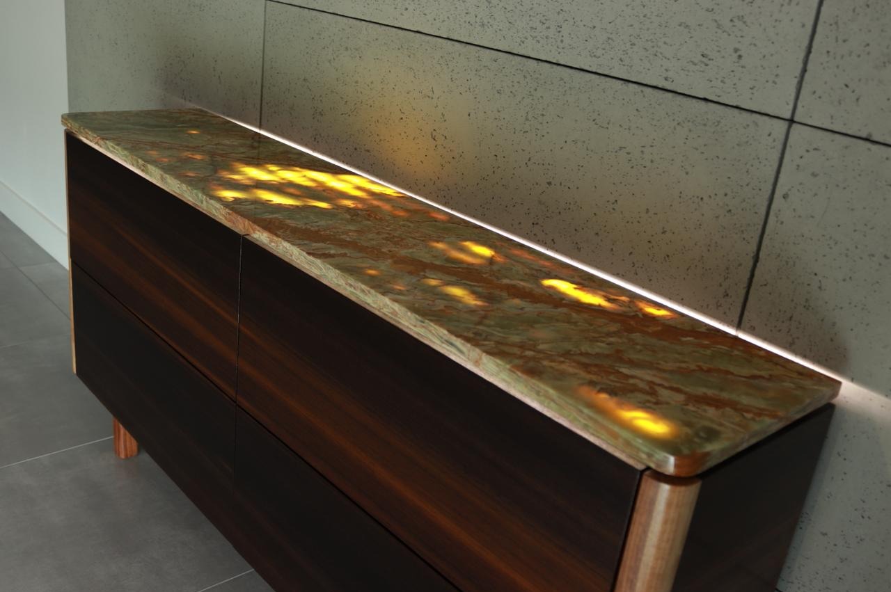Komoda z ciemnego drewna z podświetlanym blatem z zielono-brązowego kamienia na tle szarej ściany z płytek.