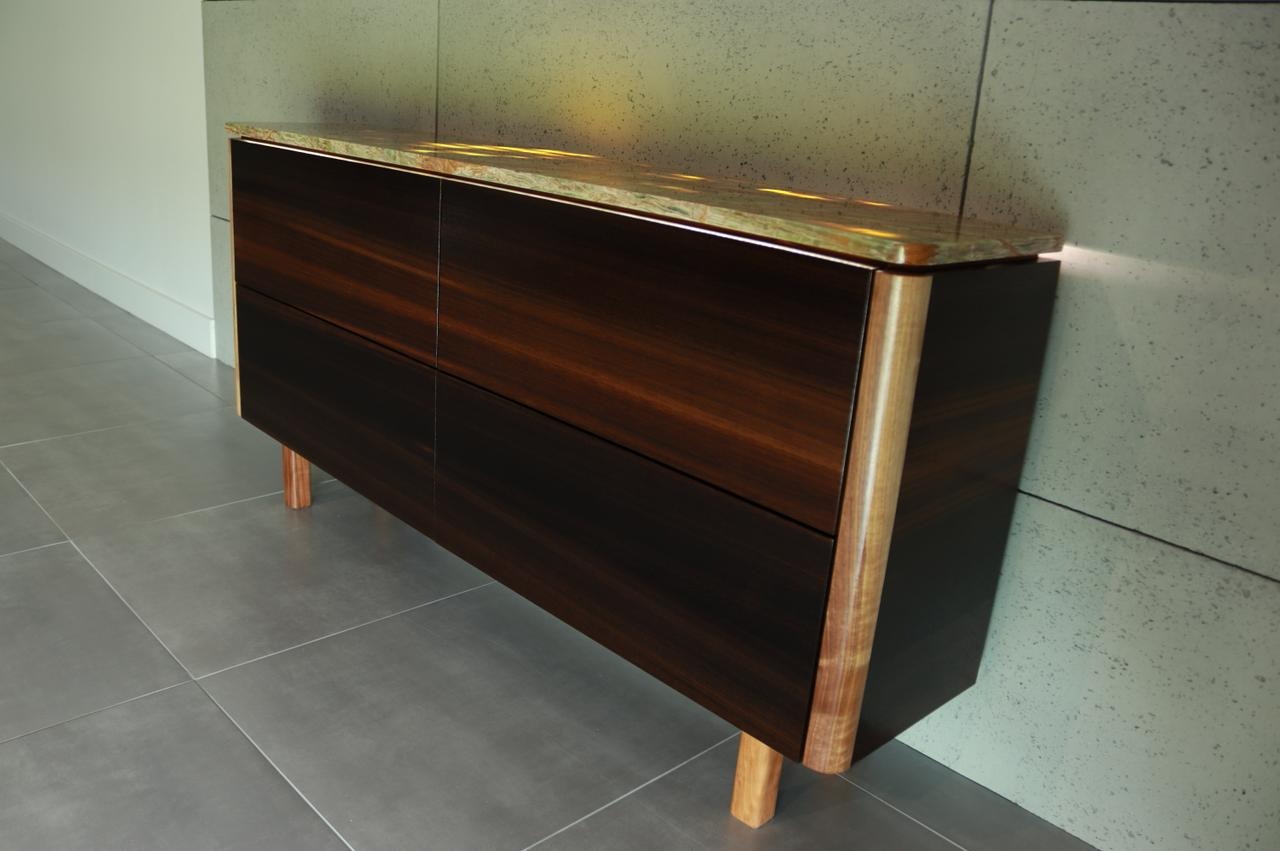 Nowoczesna komoda z ciemnego drewna z blatem z kamienia naturalnego, ustawiona na szarych płytkach podłogowych, z widocznymi elementami wykończeniowymi z jasnego drewna.