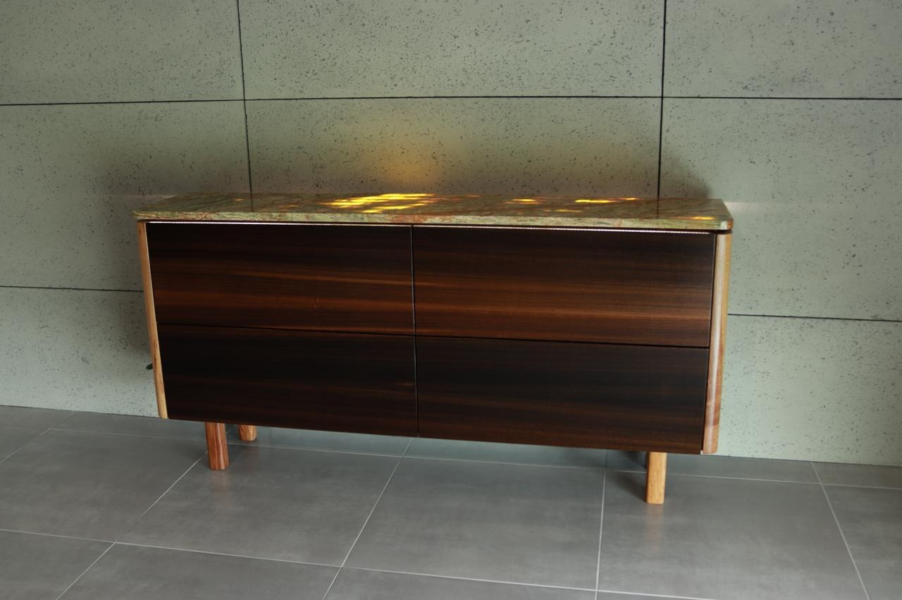 Nowoczesna komoda z ciemnego drewna z blatem z podświetlonego onyksu, na tle ściany z szarych płyt i szarej podłogi z płytek.