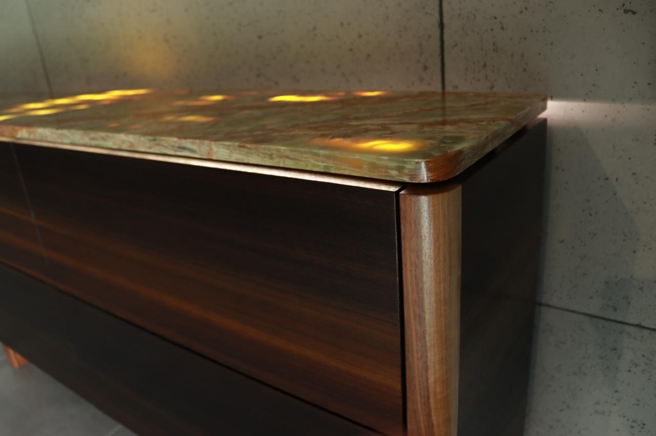 Detale wykończenia szafki z podświetlanym, marmurkowym blatem, ciemne drewno forniru i zaokrąglone, jasne nogi w minimalistycznym wnętrzu.