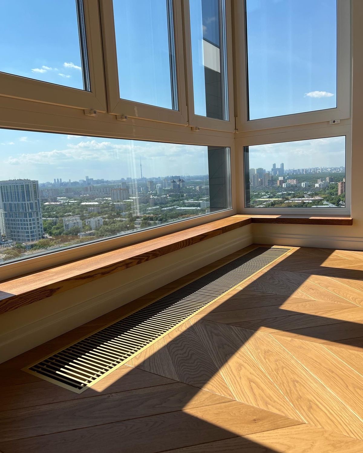 Panoramiczne okno narożne z widokiem na miasto, drewniany parapet i parkiet w jodełkę, promienie słoneczne na podłodze.