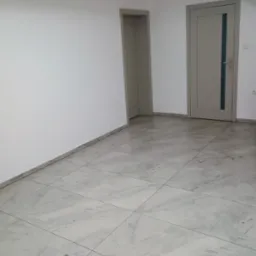 Jasne, przestronne wnętrze z podłogą wyłożoną marmurkowymi płytkami, świeżo pomalowanymi białymi ścianami i dwiema szarymi ościeżnicami z drzwiami w tle, ukazujące minimalistyczny styl wykończenia.