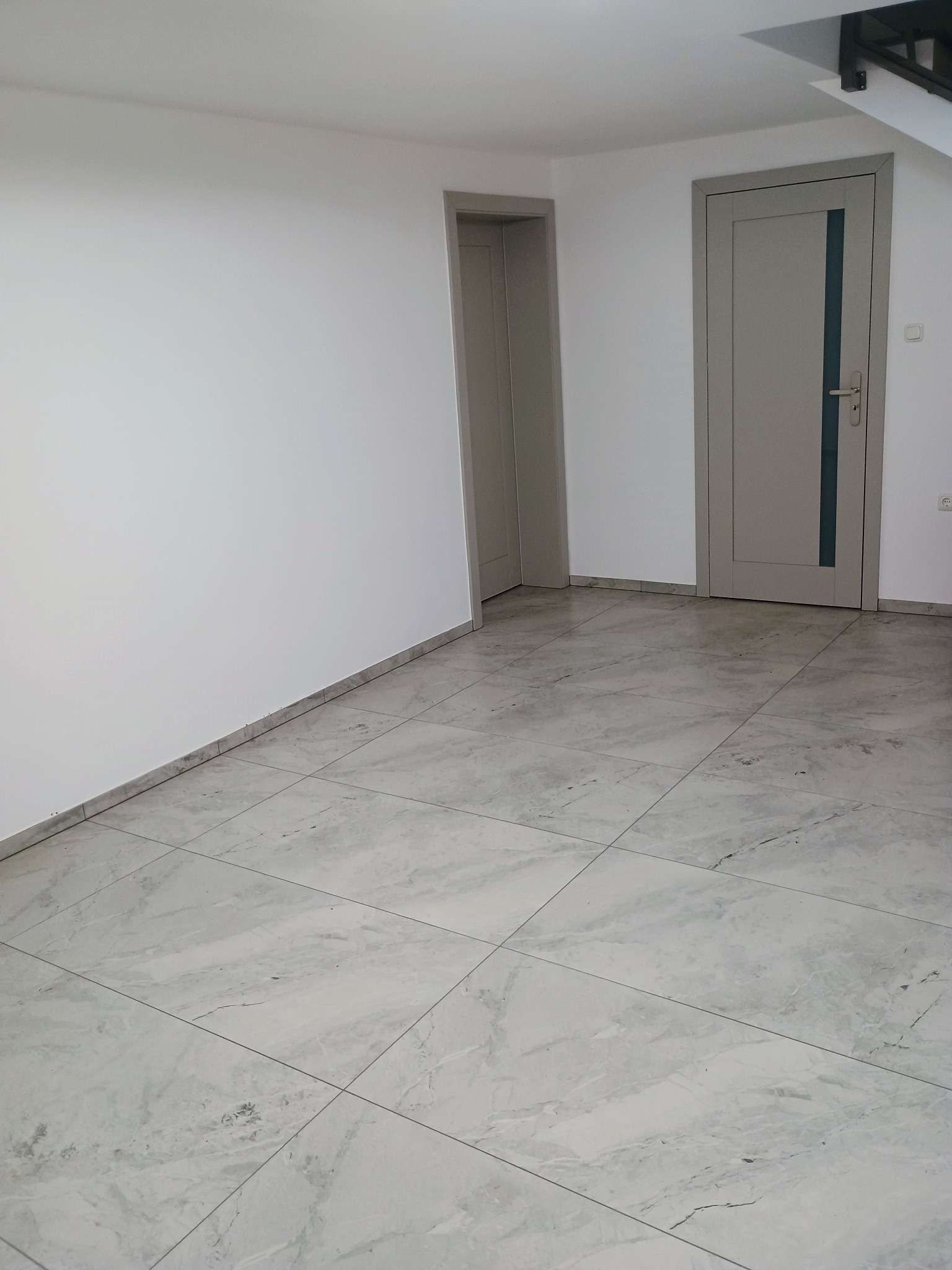 Jasne, przestronne wnętrze z podłogą wyłożoną marmurkowymi płytkami, świeżo pomalowanymi białymi ścianami i dwiema szarymi ościeżnicami z drzwiami w tle, ukazujące minimalistyczny styl wykończenia.