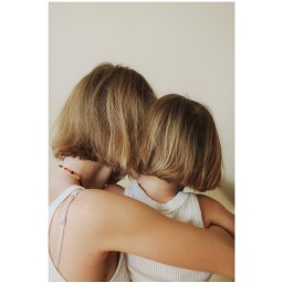 Ania Smol - Matka obejmuje córkę od tyłu, obie mają krótkie blond włosy i białe ubrania, widoczny naszyjnik z bursztynu na szyi matki.