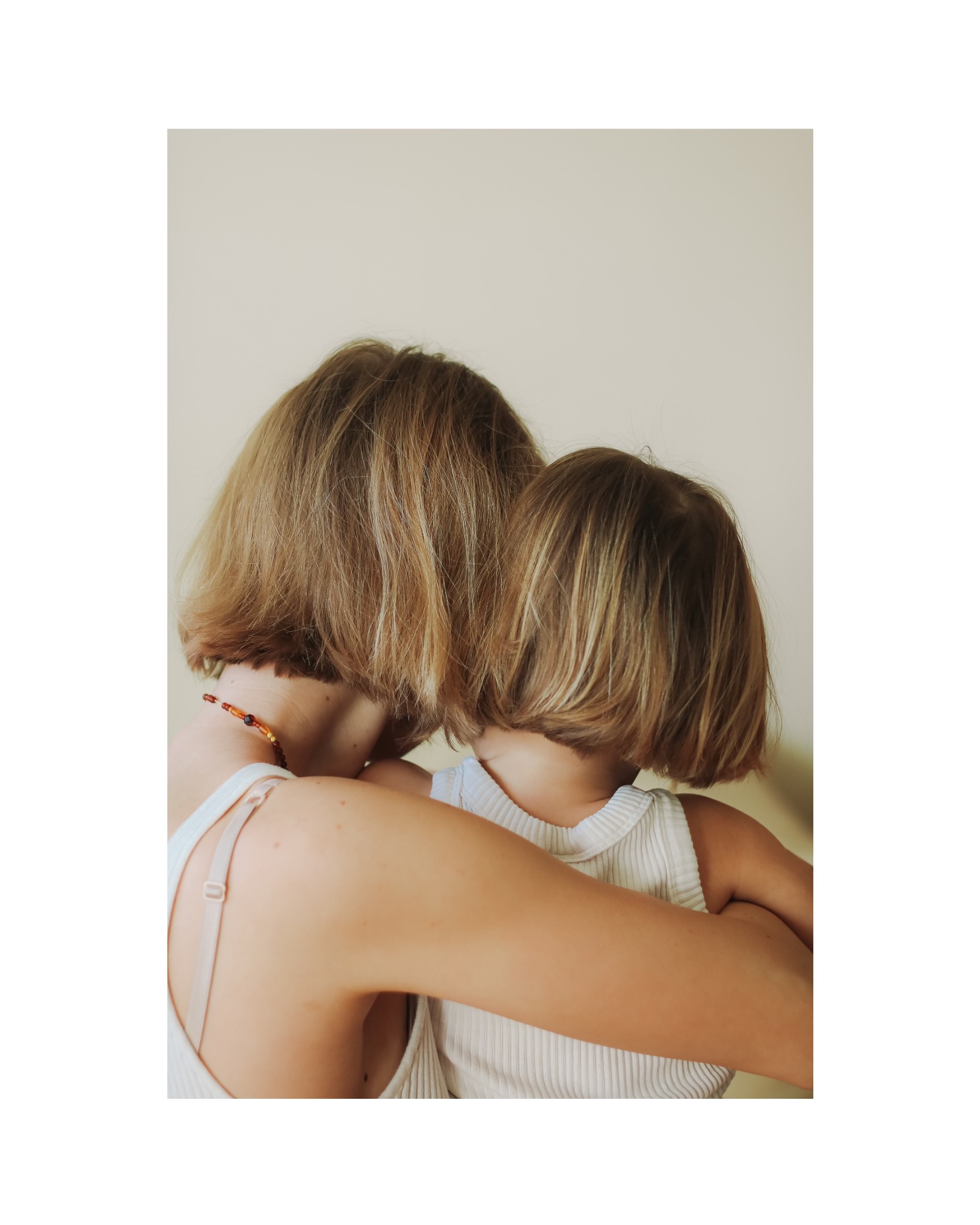 Matka obejmuje córkę od tyłu, obie mają krótkie blond włosy i białe ubrania, widoczny naszyjnik z bursztynu na szyi matki.