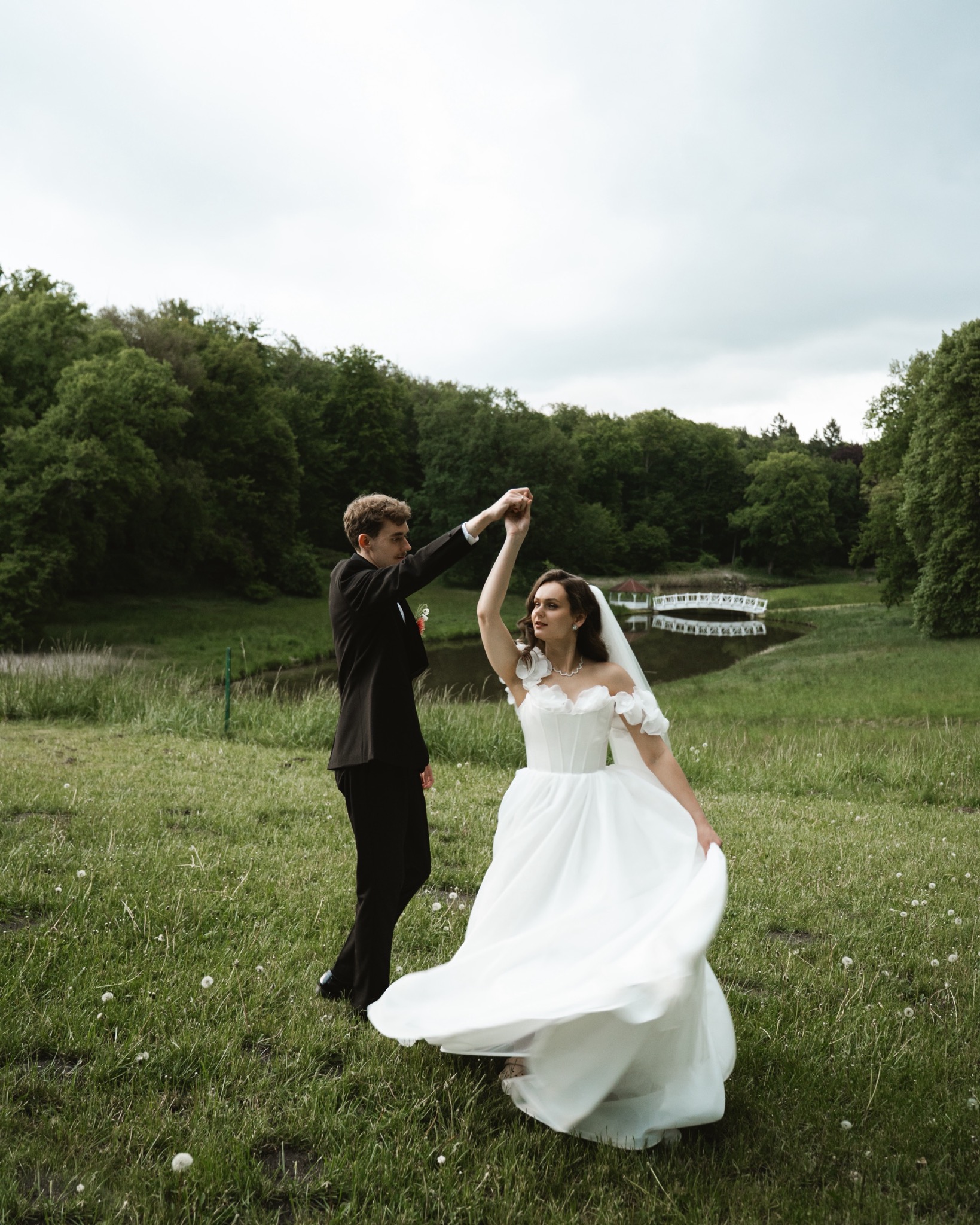 Para młoda tańczy na zielonej łące, w tle drzewa i mały mostek nad wodą. Pan młody w czarnym smokingu, panna młoda w białej sukni z welonem.