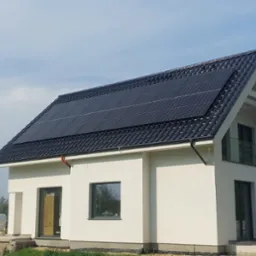 Nowoczesny dom jednorodzinny z czarną dachówką i panelami słonecznymi na dachu, widziany z poziomu gruntu przy słonecznej pogodzie, z widocznym samochodem dostawczym w tle.