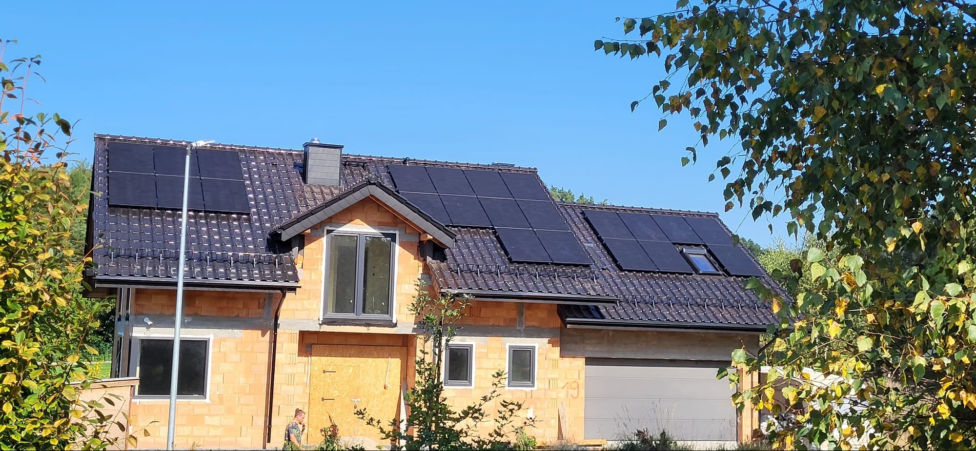 Dom w trakcie budowy z zamontowanymi panelami słonecznymi na dachu pokrytym ciemną dachówką, widoczne okna i brama garażowa.