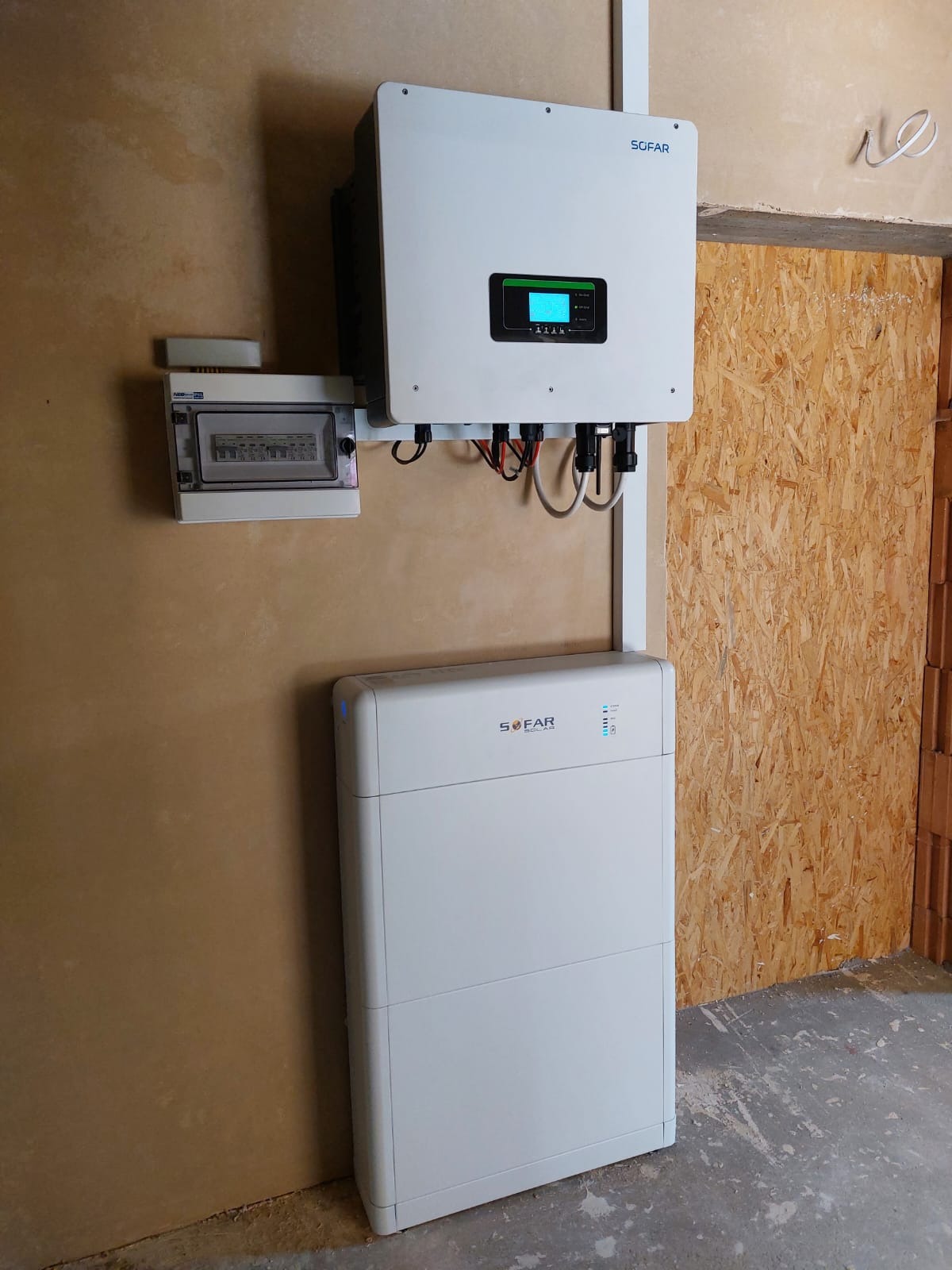 Biały inwerter i akumulator energii marki Sofar zamontowane na ścianie w surowym wnętrzu, obok skrzynka elektryczna i fragment ściany z płyt OSB.