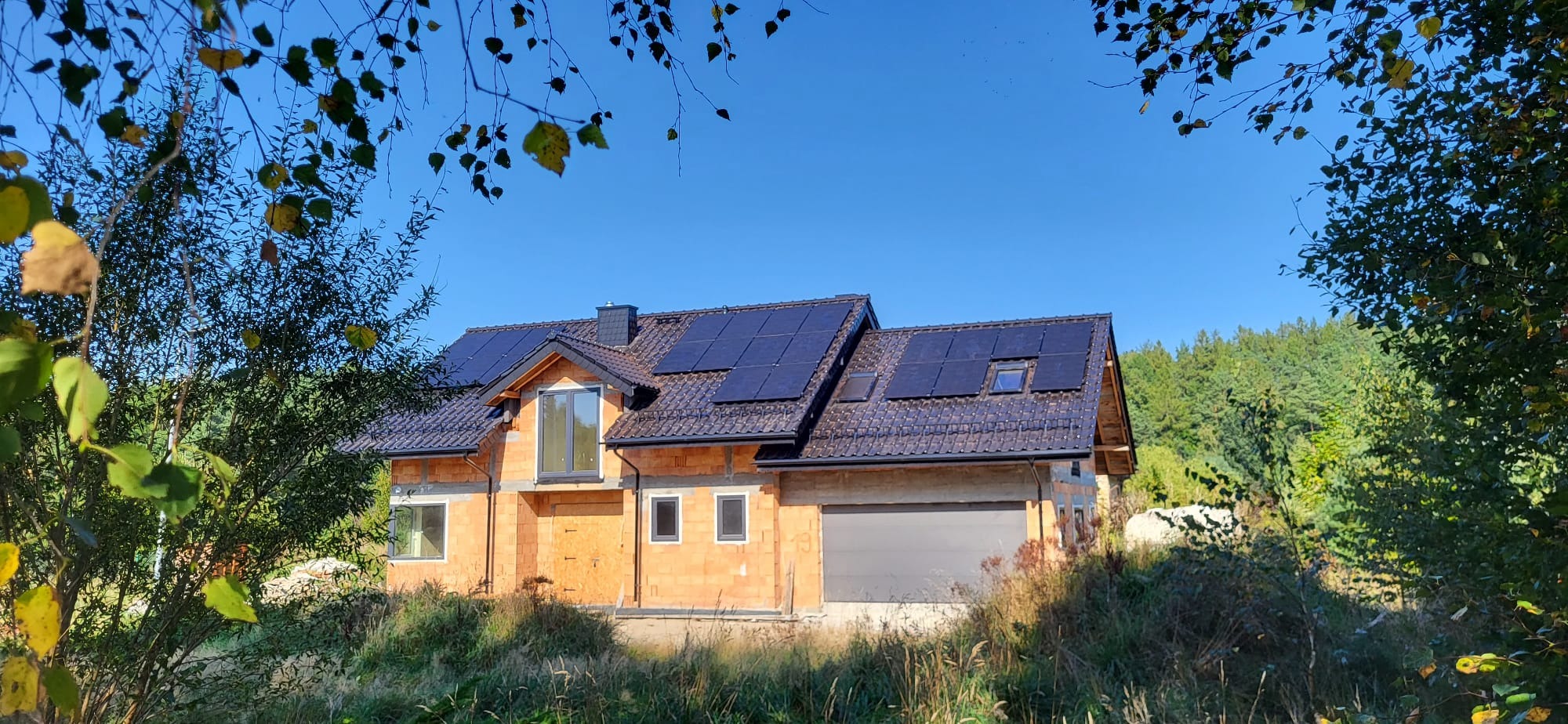 Dom jednorodzinny w trakcie budowy, z zamontowanymi panelami słonecznymi na dachu, widok z poziomu gruntu, z fragmentami drzew na pierwszym planie.