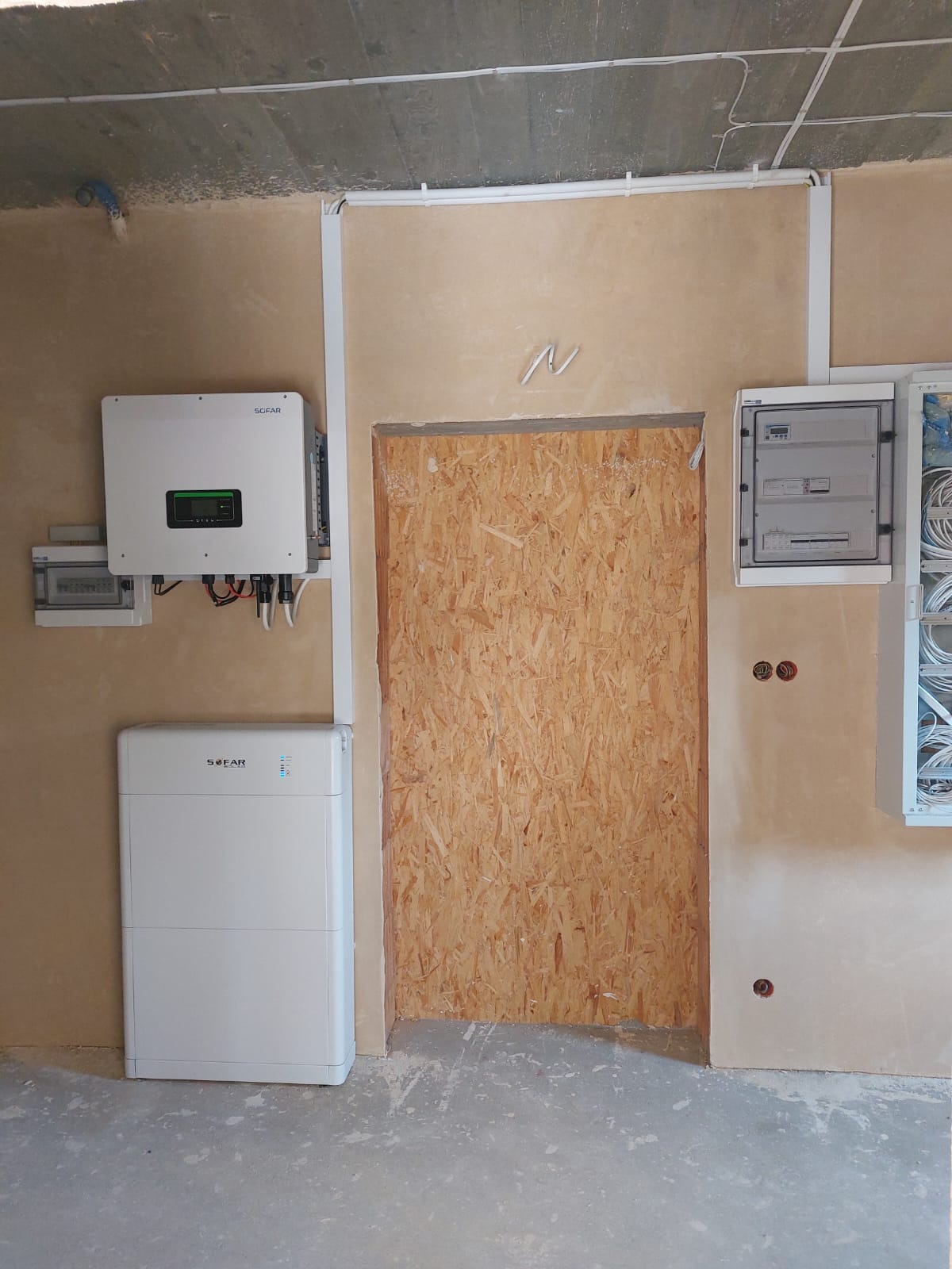Instalacja fotowoltaiczna w surowym wnętrzu: falownik i akumulator energii Sofar zamontowane na ścianie obok skrzynki elektrycznej, z zamurowanym otworem drzwiowym w tle.