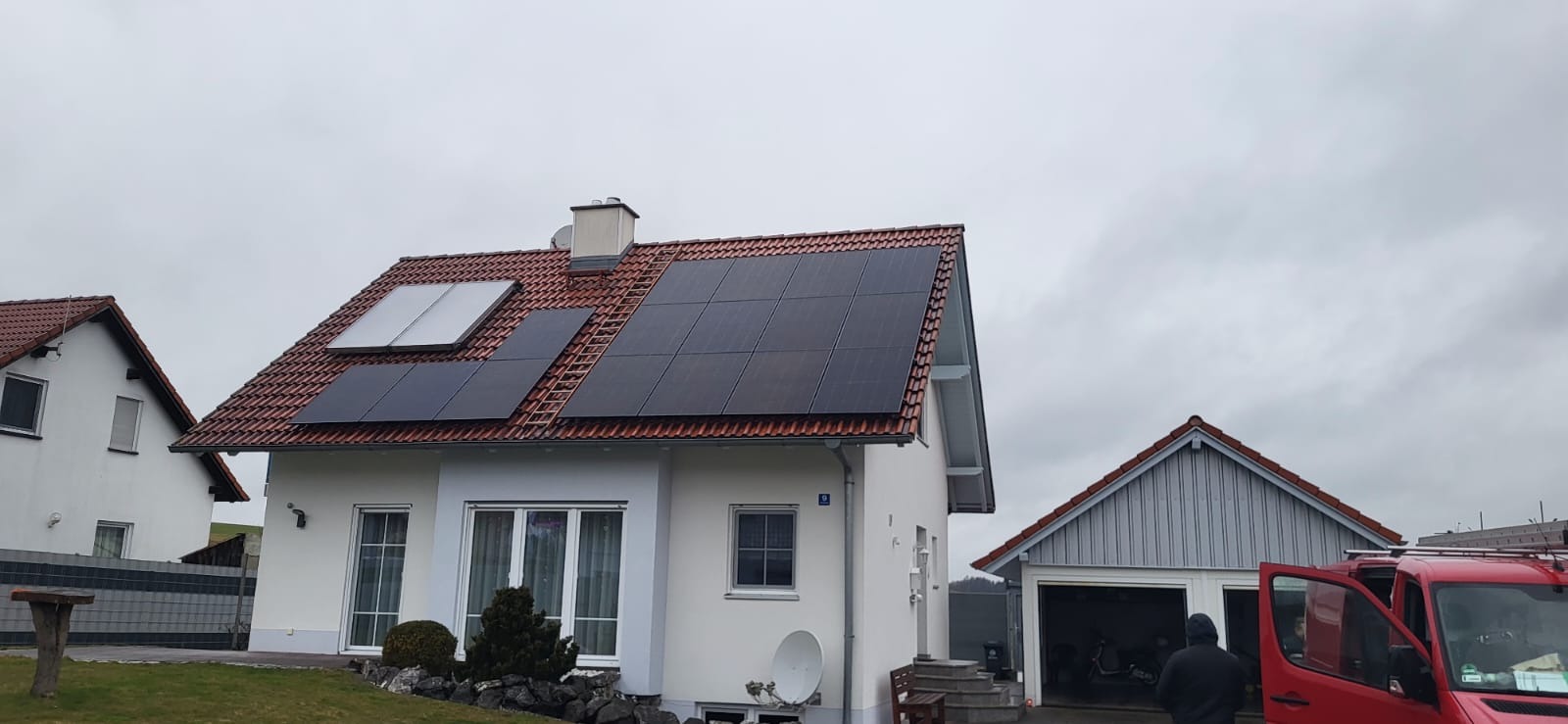 Dom jednorodzinny z panelami słonecznymi na dachu, widoczny z perspektywy ulicy, z czerwonym samochodem serwisowym w tle.