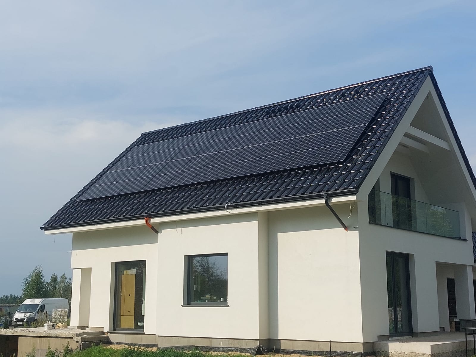 Nowoczesny dom jednorodzinny z czarną dachówką i panelami słonecznymi na dachu, widziany z poziomu gruntu przy słonecznej pogodzie, z widocznym samochodem dostawczym w tle.