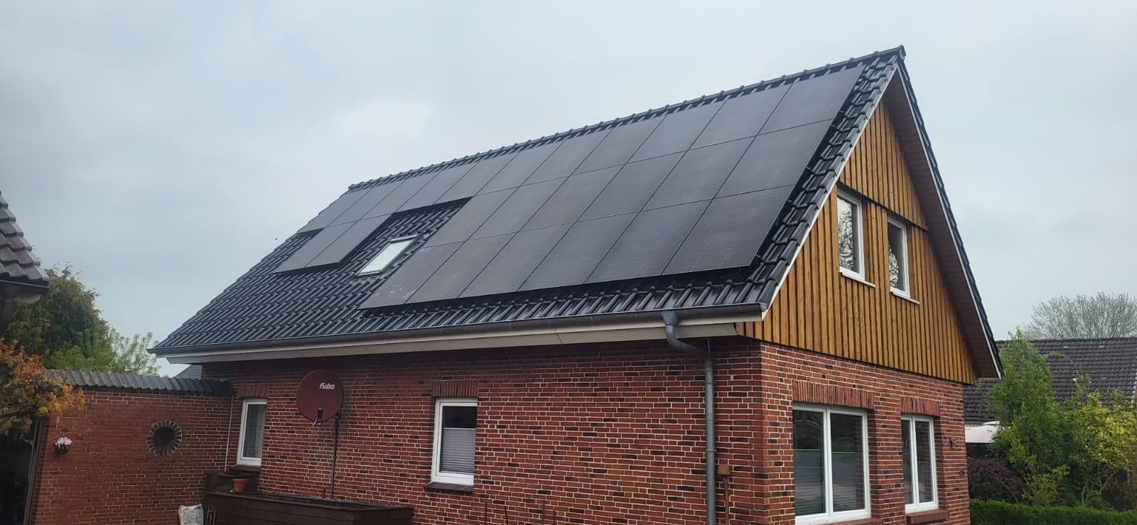 Czerwony ceglany dom jednorodzinny z panelami słonecznymi na dachu, widoczne okno dachowe, drewniana elewacja szczytowa i antena satelitarna.