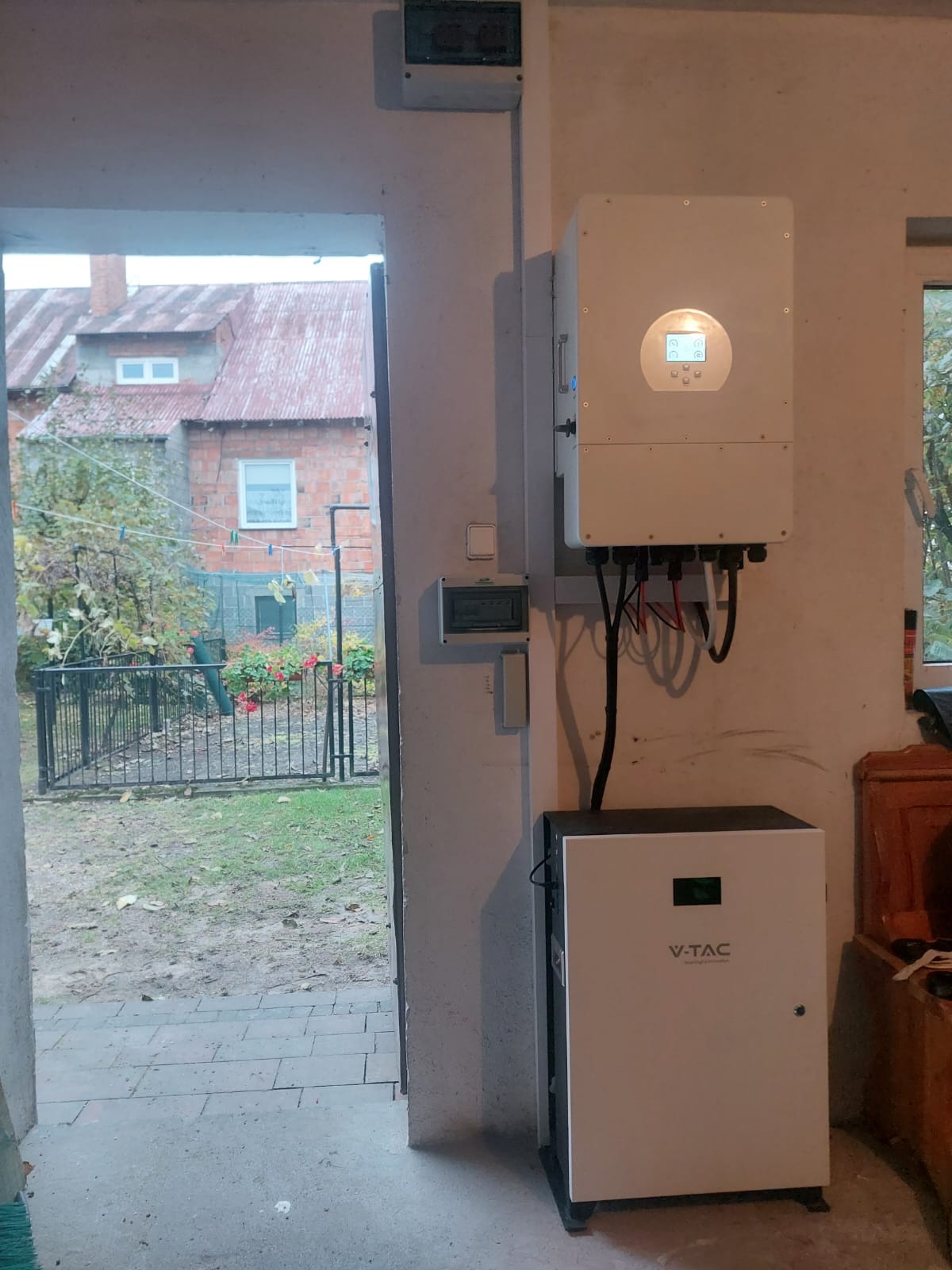 Wnętrze garażu z zainstalowanym inwerterem i magazynem energii V-TAC na tle otwartych drzwi z widokiem na ogród z suszącym się praniem.